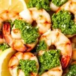 Grilled shrimp skewers brushed with vibrant green pesto, slightly charred, served on a plate with lemon wedges and sprinkled with Parmesan