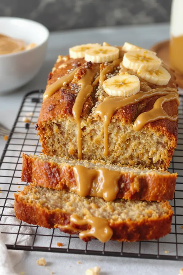 A freshly baked loaf of gluten-free banana bread on a cooling rack, with a golden-brown crust, sliced to reveal a moist, nut-studded interior. Topped with banana slices, almond butter drizzle, and chia seeds on a modern kitchen counter