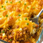 A golden, bubbly ground beef casserole in a 9×13-inch baking dish, topped with crispy hash browns and melted cheddar cheese, with a sprinkle of green onions and a serving spoon ready on the side