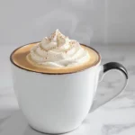 A steaming pumpkin spice latte in a ceramic mug, topped with whipped cream and a dusting of cinnamon all on a modern kitchen counter