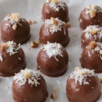 Small round keto fat bombs with a smooth chocolate finish, topped with a sprinkle of shredded coconut and chopped nuts, arranged neatly on a parchment‑lined plate