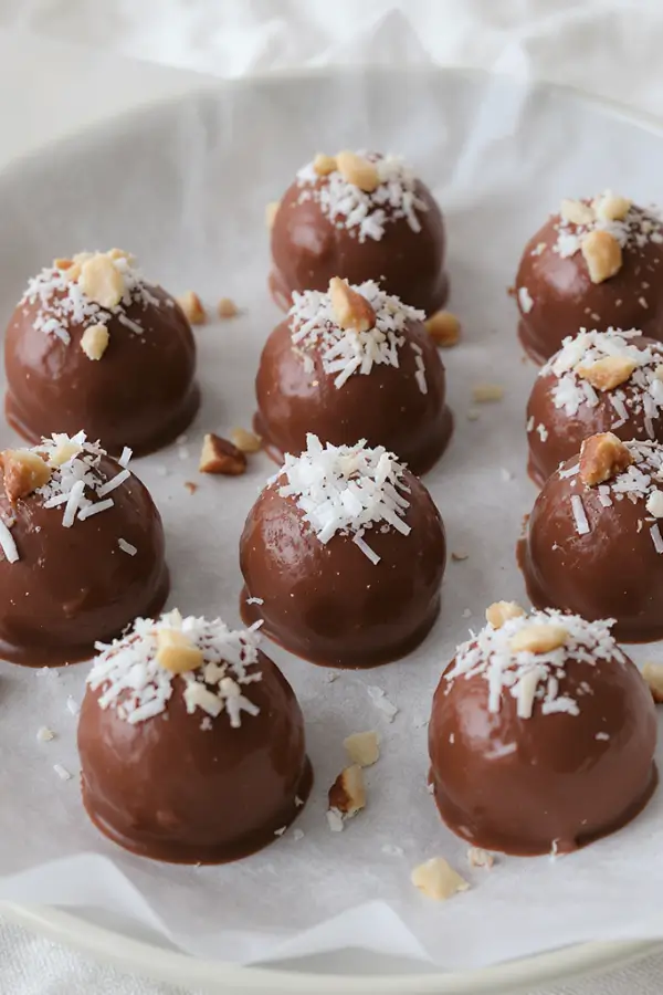 Small round keto fat bombs with a smooth chocolate finish, topped with a sprinkle of shredded coconut and chopped nuts, arranged neatly on a parchment‑lined plate