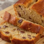 A golden loaf of banana bread on a wooden board, sliced to reveal a moist interior with melted chocolate chips and crunchy walnut pieces