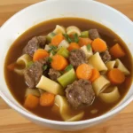 A bowl of crockpot beef and noodles, featuring tender beef chunks, wide egg noodles, carrots, and celery in a rich, savory broth set on a wooden table