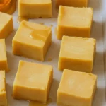 A tray of creamy maple fudge squares, golden-brown with a smooth finish, arranged neatly on parchment paper with a drizzle of maple syrup nearby