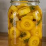 one Glass jar filled with vibrant yellow banana pepper rings in golden brine, speckled with mustard seeds, black peppercorns, garlic slices, and sprigs of dill, sitting on a kitchen counter