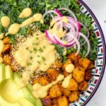 Colorful vegan harvest bowl with fluffy quinoa, roasted sweet potatoes and crispy chickpeas, vibrant massaged kale, sliced avocado, and thin red onion slices, drizzled with creamy Dijon mustard dressing served in vibrant bowls
