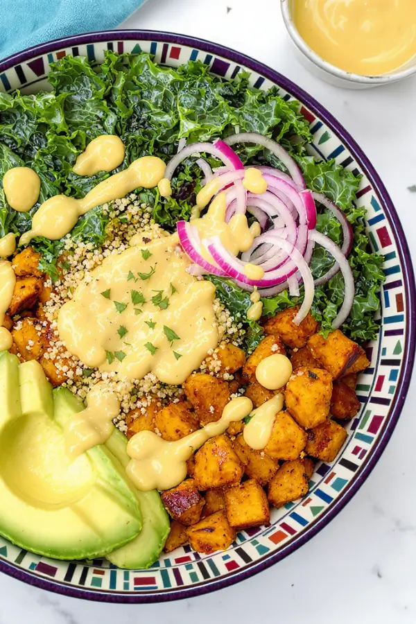 Colorful vegan harvest bowl with fluffy quinoa, roasted sweet potatoes and crispy chickpeas, vibrant massaged kale, sliced avocado, and thin red onion slices, drizzled with creamy Dijon mustard dressing served in vibrant bowls