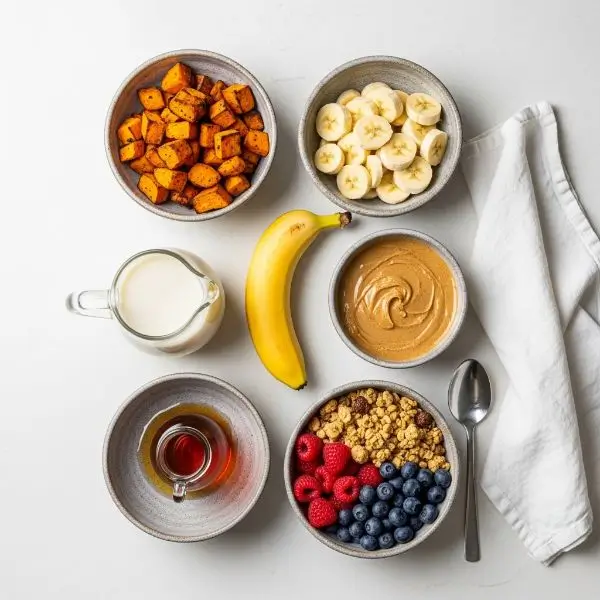 Smoothie ingredients in ceramic bowls including roasted sweet potato, banana, almond milk, nut butter, maple syrup, and toppings like granola and berries on a white countertop