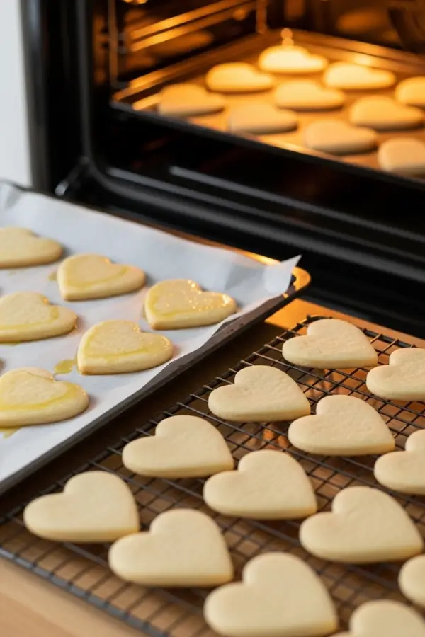 Freshly baked heart-shaped sugar cookies on parchment-lined baking sheets, lightly golden edges, cookies cooling on a wire rack