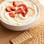 A white ceramic serving bowl filled with fluffy no bake cheesecake dip, swirled on top, garnished with fresh sliced strawberries