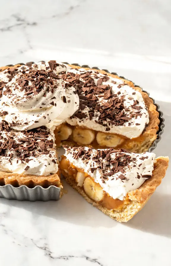 Whole banoffee pie with whipped cream and dark chocolate shavings on top showing three distinct layers of biscuit crust golden toffee and banana