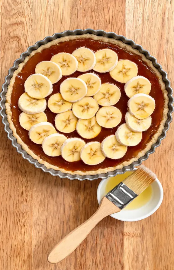 Ripe banana slices arranged in an overlapping layer over golden set toffee inside a banoffee pie crust with a small bowl of lemon juice nearby