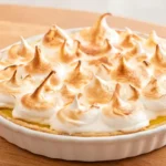 A whole golden lemon meringue pie on a white ceramic pie dish, set on a wooden table, with tall billowy toasted meringue peaks in golden and white
