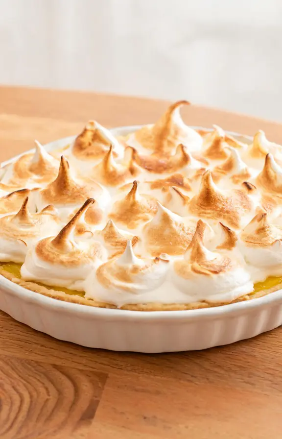 A whole golden lemon meringue pie on a white ceramic pie dish, set on a wooden table, with tall billowy toasted meringue peaks in golden and white