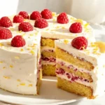 Finished lemon raspberry layer cake, sliced to show the interior, fresh raspberries on top, golden lemon zest scattered, cream cheese frosting