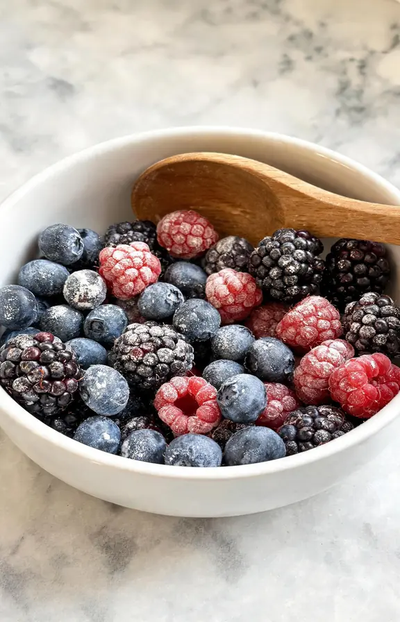 A bowl of fresh mixed berries, blueberries, raspberries and blackberries, dusted lightly with cornstarch, wooden spoon resting in the bowl