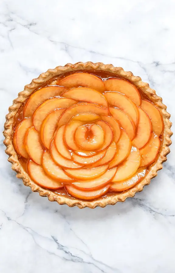 Overhead view of a glazed fresh peach tart with concentric golden peach slices and shiny apricot glaze on a white marble surface