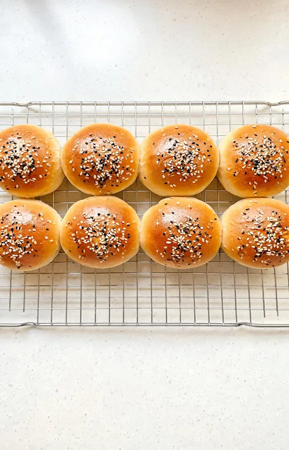 Eight golden soft sourdough discard hamburger buns cooling on a wire rack, topped with sesame seeds and egg wash shine