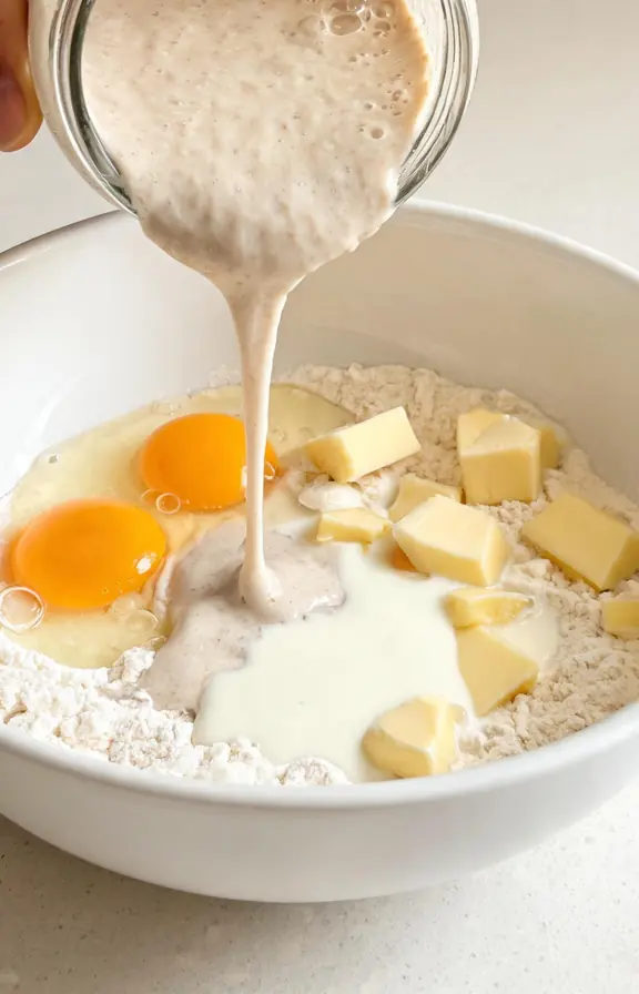 Sourdough discard being poured from a jar into a bowl of enriched dough ingredients including eggs, butter, and milk