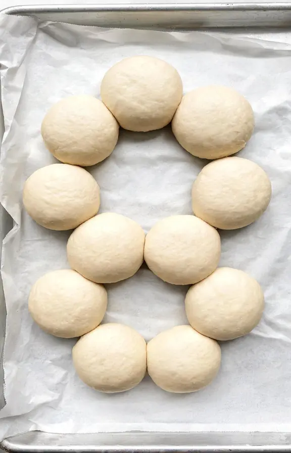 Eight shaped sourdough discard hamburger buns on a parchment-lined baking sheet before proofing, evenly spaced