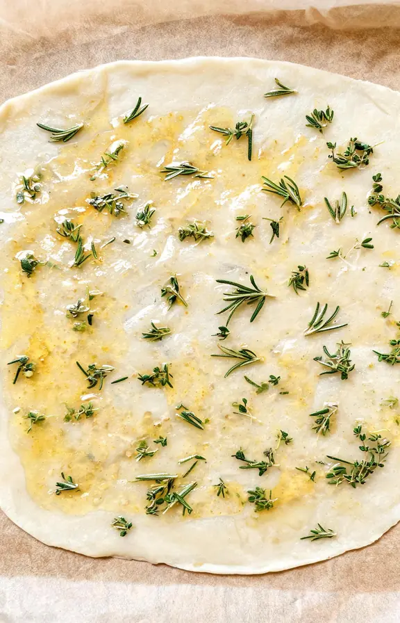 Thin translucent dough rolled on parchment, brushed with garlic oil, scattered with rosemary and thyme leaves