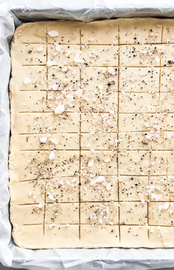Scored dough sheet on parchment, flaky sea salt and cracked pepper visible