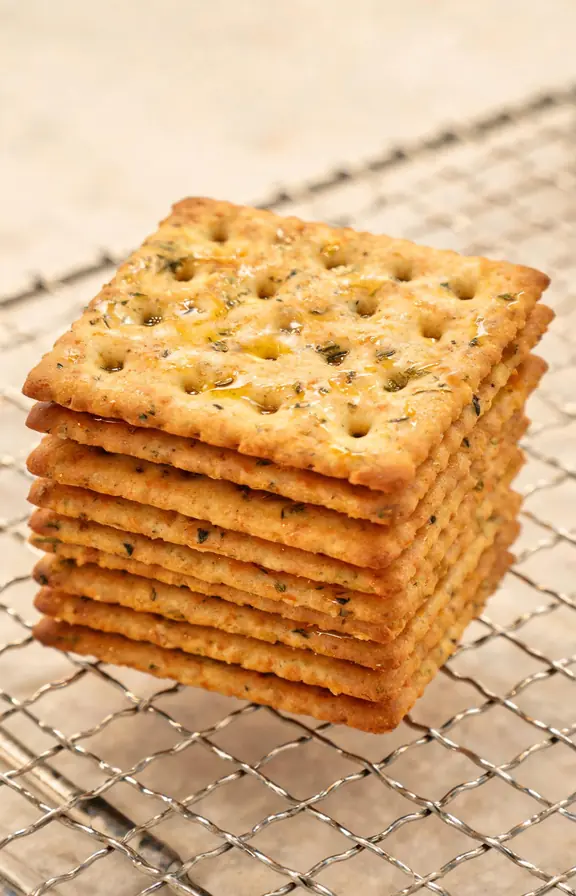Stack of finished crackers on wire cooling rack, dimpled surface, olive oil sheen, dried herb specks