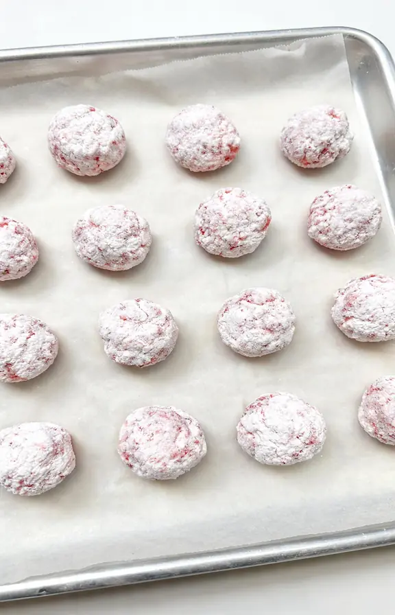 Powdered sugar-coated strawberry crinkle cookie dough balls on a parchment-lined baking sheet