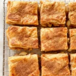 Golden apple pie bars made with puff pastry, cut into squares and cooling on a wire rack with a dusting of cinnamon sugar on top
