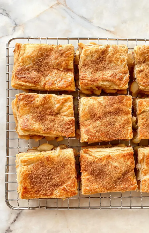 Golden apple pie bars made with puff pastry, cut into squares and cooling on a wire rack with a dusting of cinnamon sugar on top