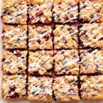 Sixteen glossy cherry pie bars cut into squares on a parchment lined board, showing dark red cherry filling between golden shortbread layers and a drizzle of white glaze