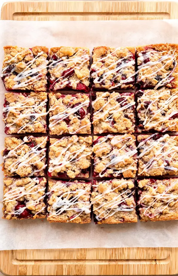 Sixteen glossy cherry pie bars cut into squares on a parchment lined board, showing dark red cherry filling between golden shortbread layers and a drizzle of white glaze