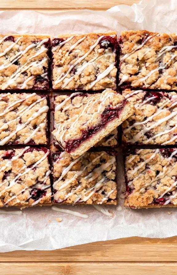 Sixteen glossy cherry pie bars cut into squares on a parchment lined board, one bar angled to show the layered cut edge