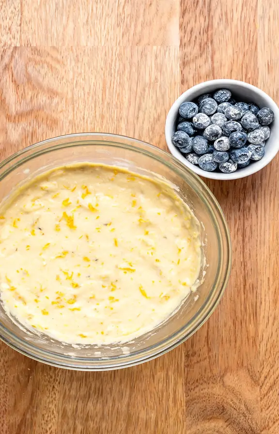 Overhead view of lemon blueberry cake batter in a glass bowl with visible lemon zest specks, next to a small bowl of flour dusted blueberries ready to be folded in