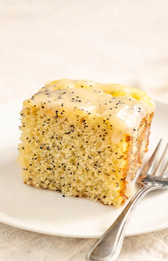Slice of lemon poppy seed cake with lemon glaze on a white plate next to whole lemons