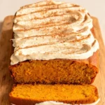 A golden-brown pumpkin loaf on a wooden board, topped with swooped cream cheese frosting