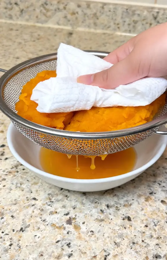 Pumpkin puree being pressed with a paper towel in a fine mesh strainer to remove excess moisture before baking
