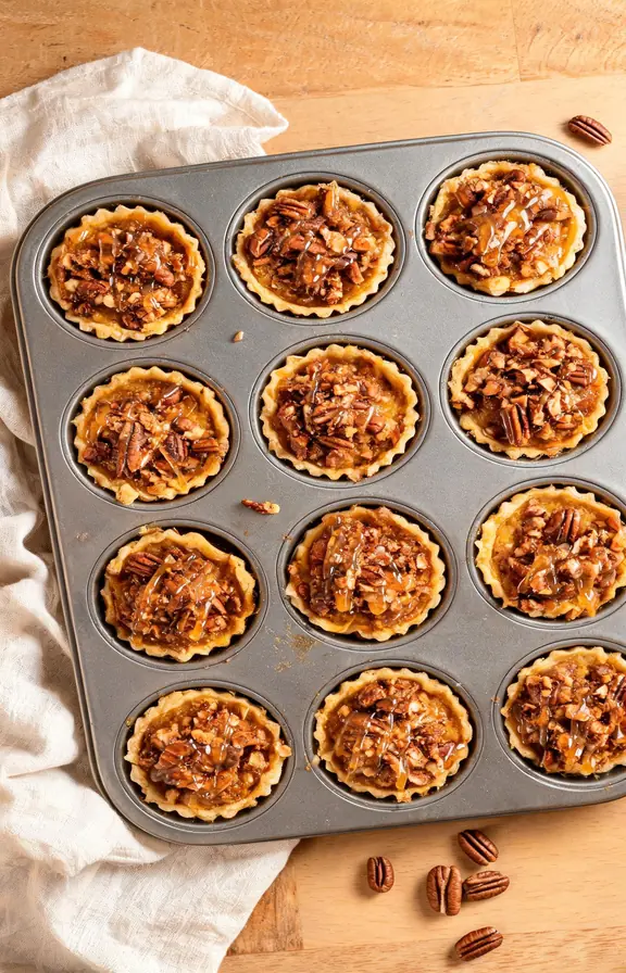 Overhead shot of pumpkin pecan tassies in a mini muffin pan topped with toasted pecan crumble and a maple glaze drizzle