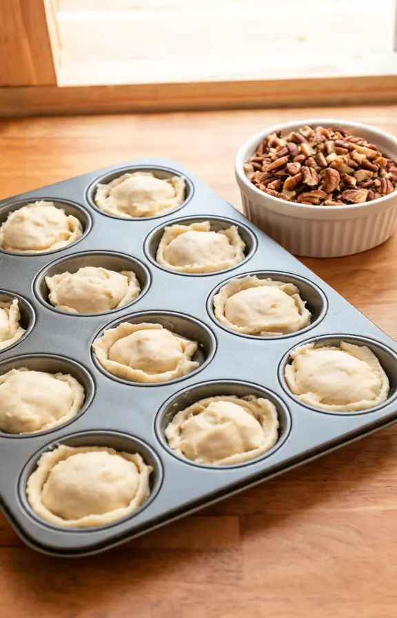 Cream cheese pastry balls pressed into a mini muffin pan chilling before baking, next to a bowl of toasted pecans