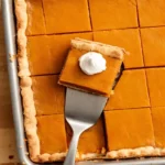 Pumpkin slab pie cut into squares on a sheet pan with a dollop of whipped cream on top