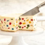A sharp knife mid-slice through a whole terrazzo cheesecake on a cake stand